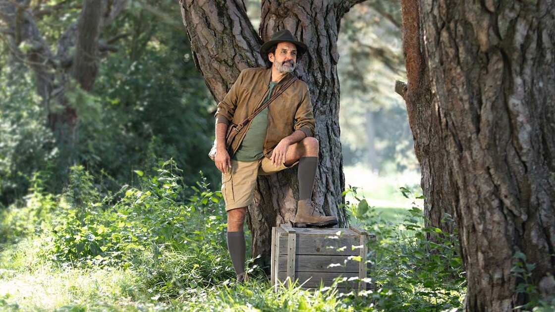 Um homem com um fato de explorador está na natureza e olha para longe. Tem calças de compressão até ao joelho.