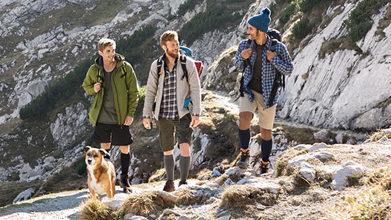 Three men with a dog in the mountains. The men are wearing compression stockings on their lower legs.